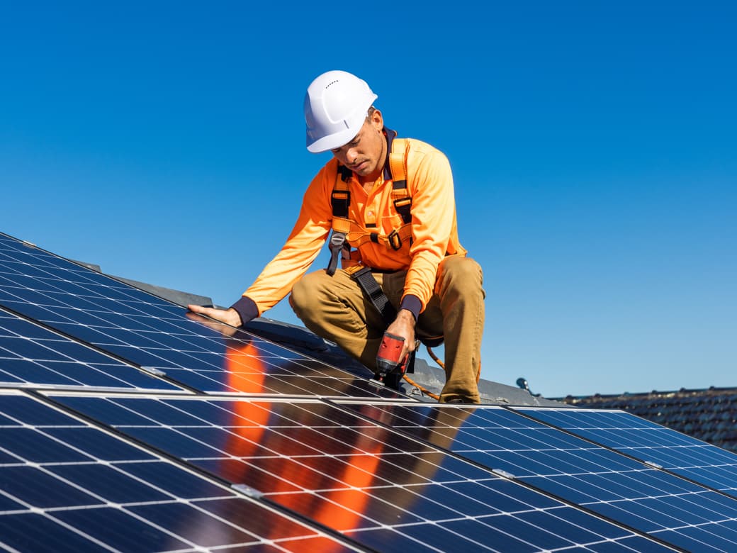 An installer in a hard hat and safety harness fixing a solar panel to a roof
